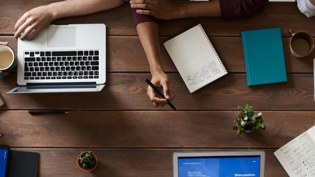 A laptop, a couple of notebooks and pen on a tabletop with some hands.